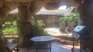 A rustic outdoor area with a stone wall, a thatched-roof hut, a grill, and a small octagonal table. - Bushmans Rock Country Lodge (Roodeplaat)