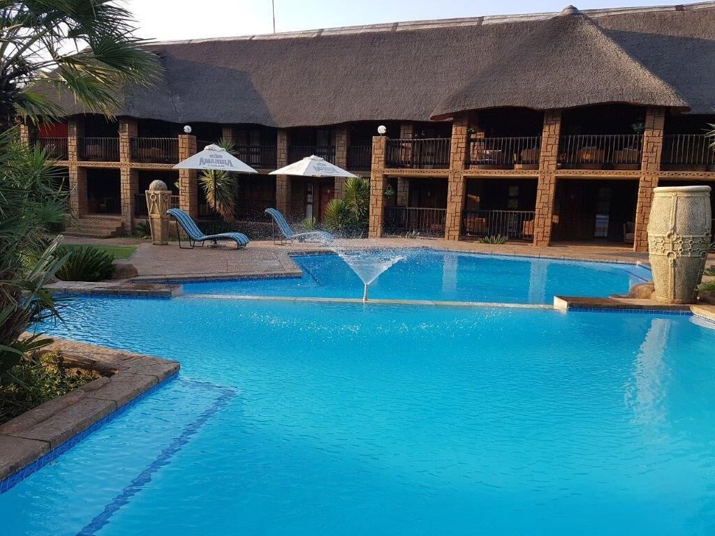 Una piscina con sillas de descanso y sombrillas, un edificio con techo de paja y balcones, y una jardinera de piedra.