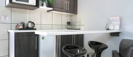 A compact kitchen with a microwave, a small refrigerator, and a coffee machine. There are two bar stools and a plant on the counter.