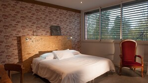 A bedroom with a brick-patterned wall, a wooden headboard, a bed with white linens, a red upholstered chair, and a window with blinds. - Le Jardin de la Reyssouze (Gorrevod)