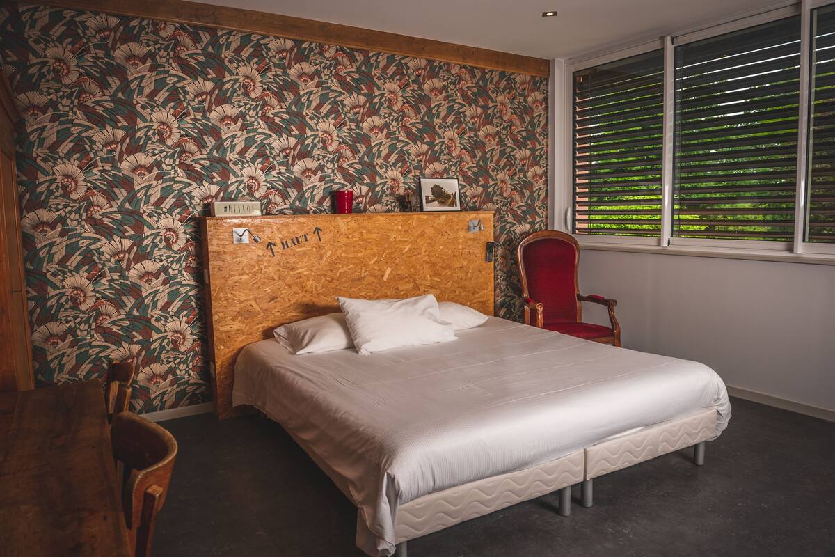 Un dormitorio con cabecera de madera, una cama con ropa de cama blanca, una silla roja y una ventana con persianas.