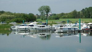 A marina with several boats docked, surrounded by greenery and a church in the background. - Le Jardin de la Reyssouze (Gorrevod)
