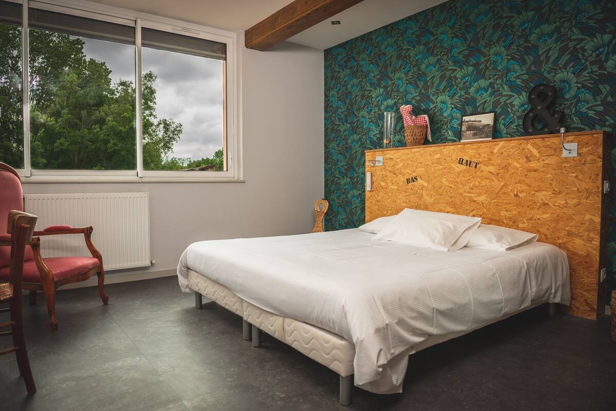 Un dormitorio con cabecera de madera, cama con ropa de cama blanca, una silla roja y una ventana con vistas a árboles.