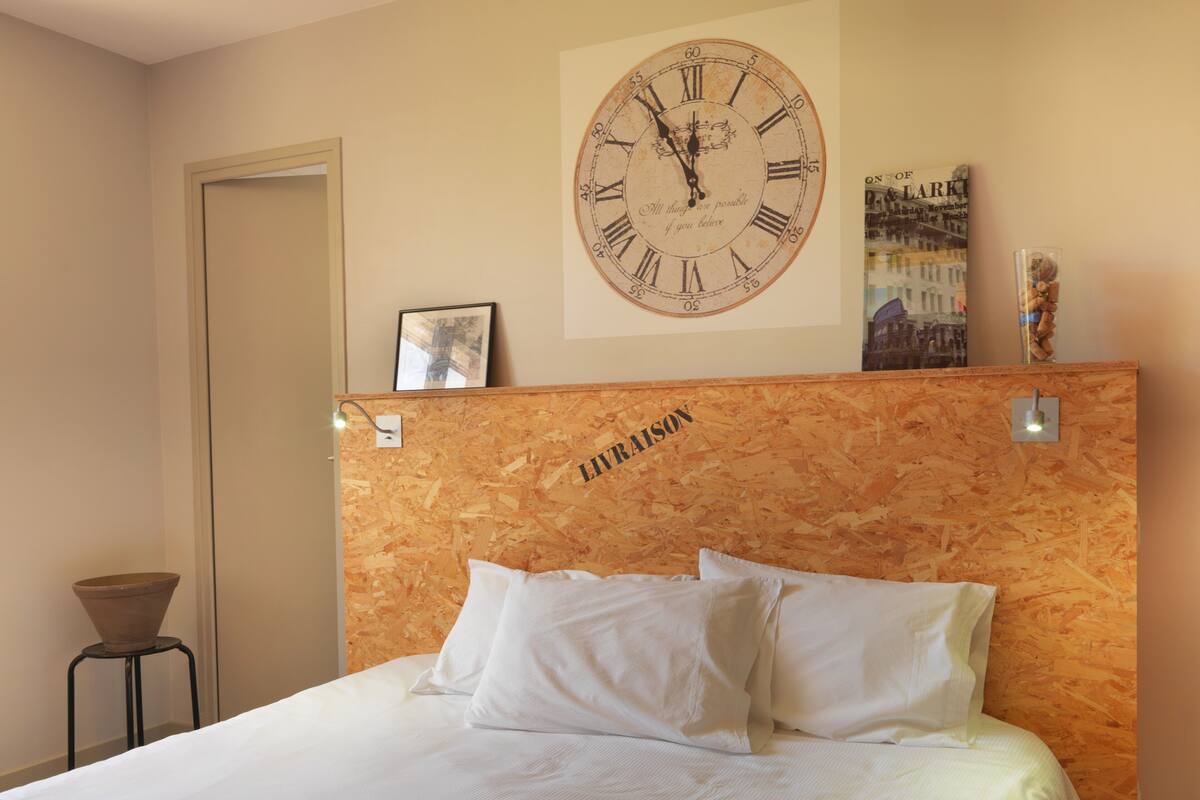 Habitación de hotel con cabecera de madera, un reloj de pared grande, un cuadro enmarcado y una mesita con un jarrón.