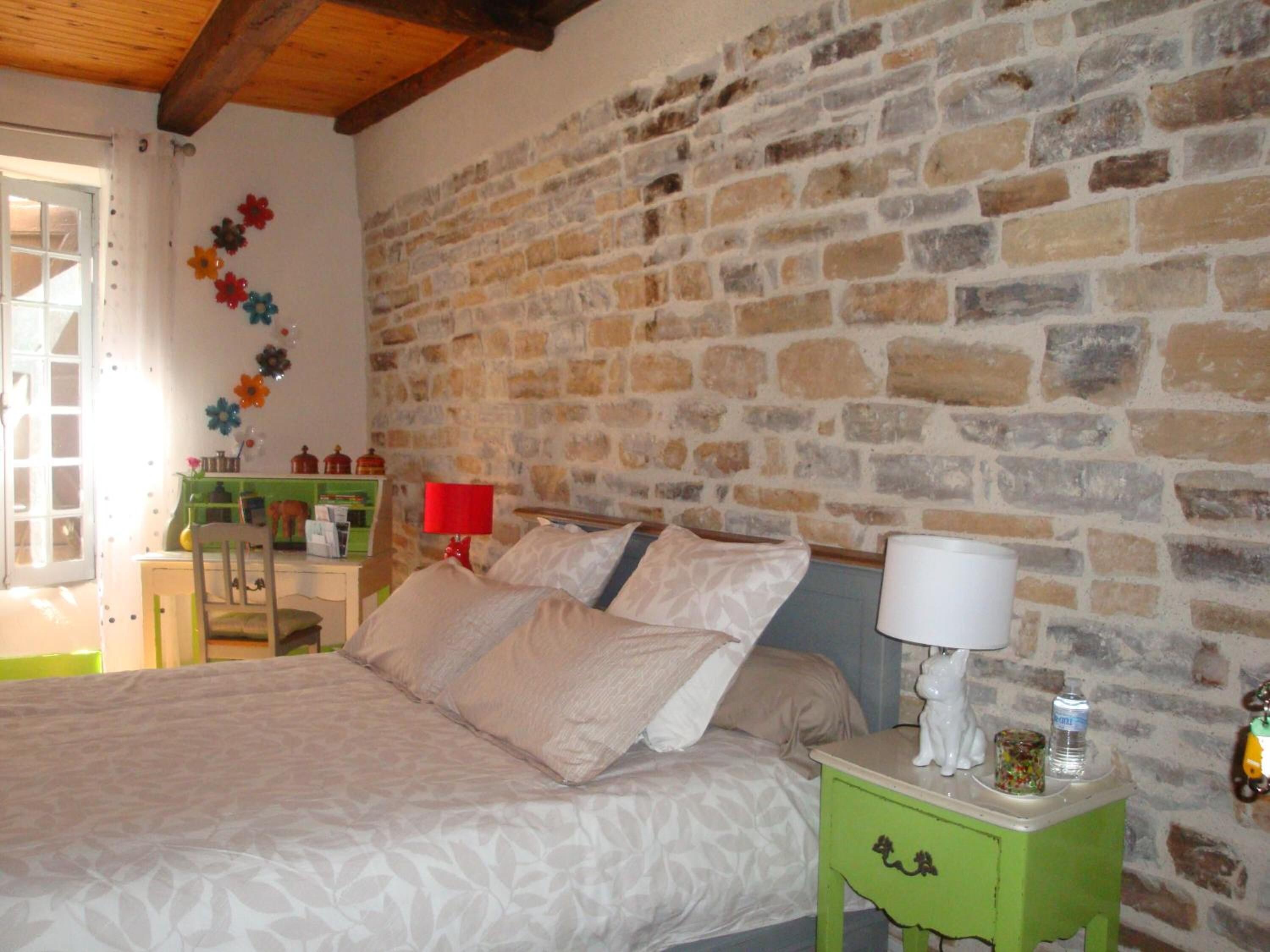 Un dormitorio con pared de piedra, una cama con ropa de cama color beige, una mesita de noche verde y una lámpara roja.
