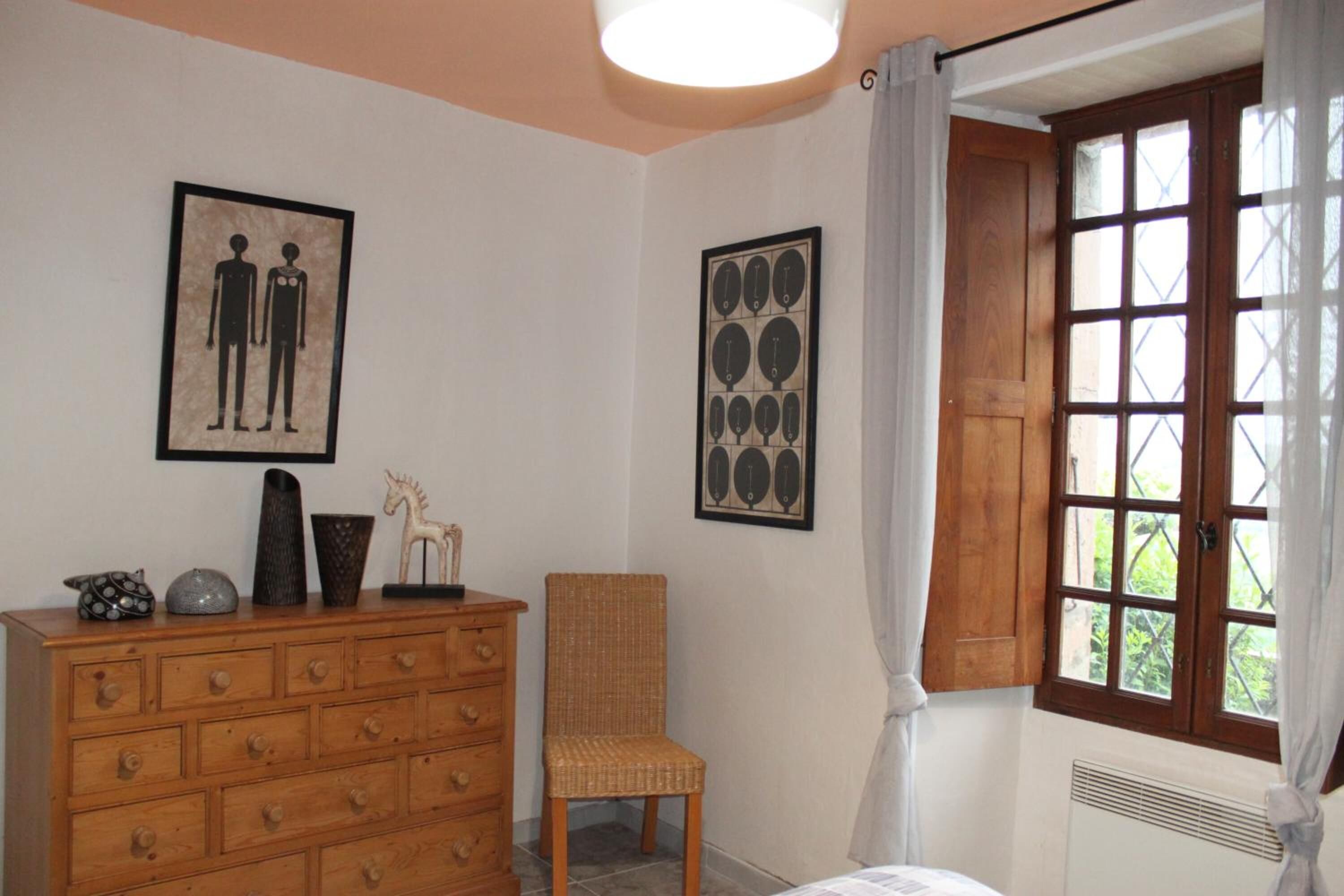 A room with a wooden dresser, a wicker chair, two framed artworks, and a window with curtains.