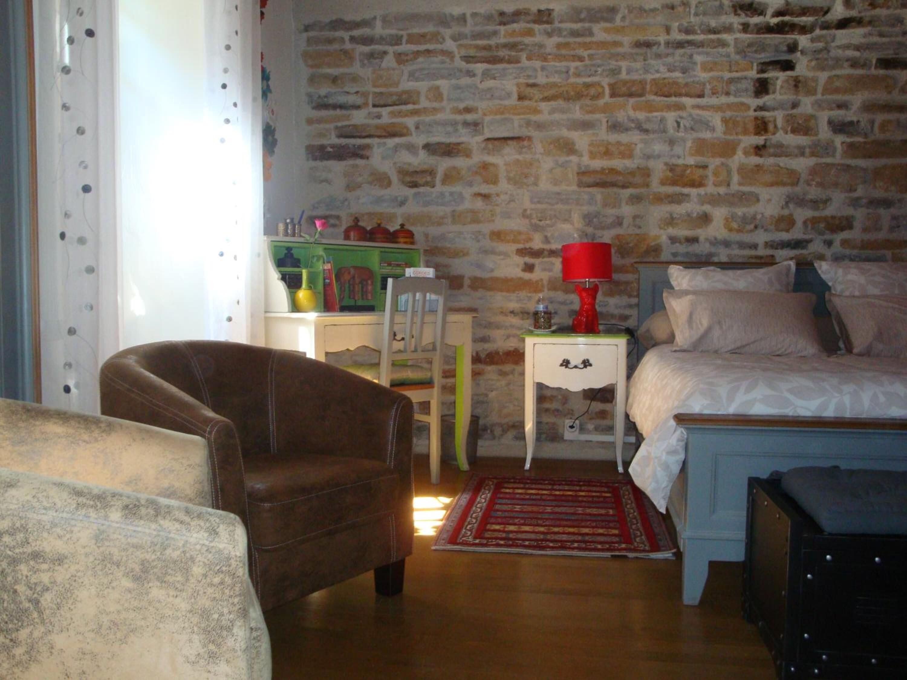 A bedroom with a stone wall, a bed with white bedding, a red lamp, and a brown leather chair.