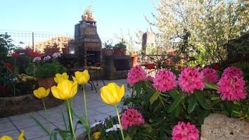 Un jardin agrémenté de fleurs jaunes et roses, une cheminée en briques et une table en métal avec des chaises.