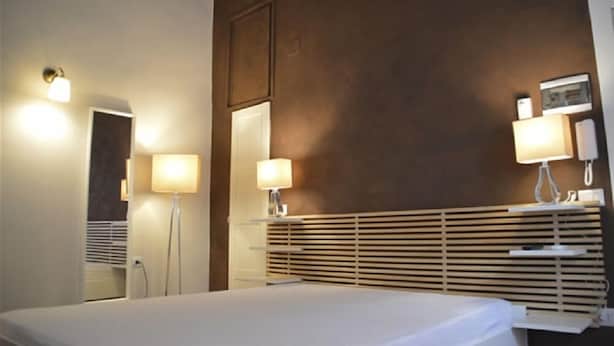 Hotellihuone, jossa on sänky, yöpöytälamput, peili ja puinen pääty.