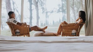 A room with a large bed, two wicker chairs, and a view of tall trees through large windows. - Andrea Lodges (Riviere des Anguilles)