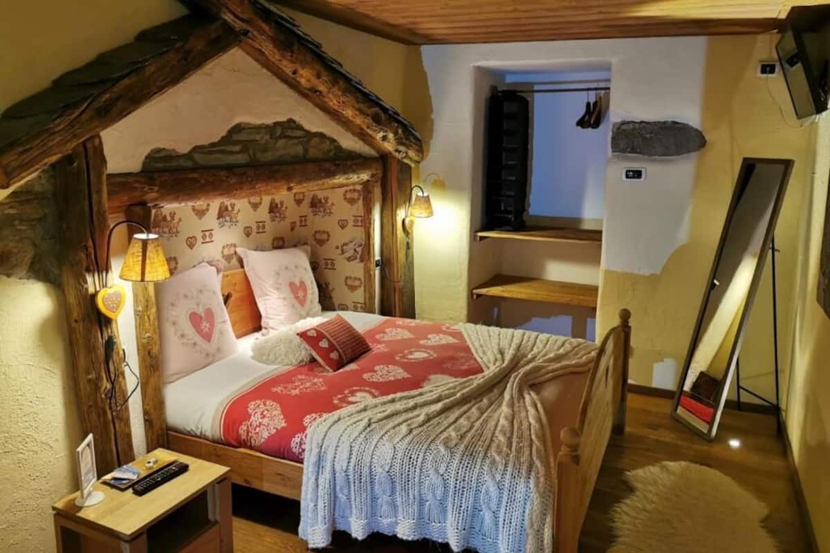 A cozy bedroom with a wooden ceiling, a bed with a red and white patterned bedspread, a small bedside table with a lamp, and a mirror leaning against the wall.