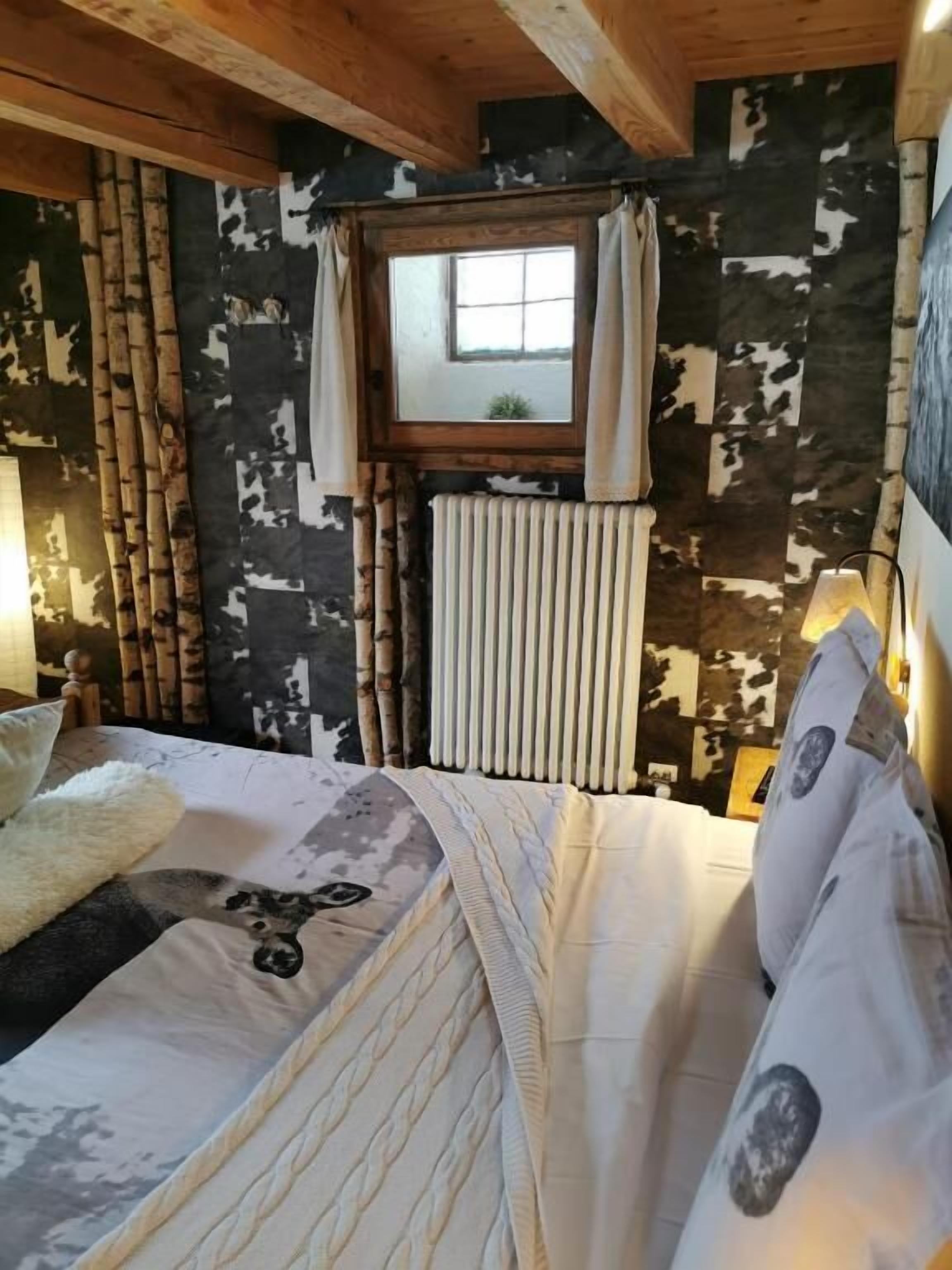 A cozy bedroom with a bed, a window with curtains, a radiator, and a wooden ceiling.