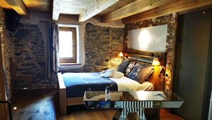 Un dormitorio con pared de piedra, vigas de madera en el techo, una cama con muchos almohadones, lámparas de noche y un espejo grande.