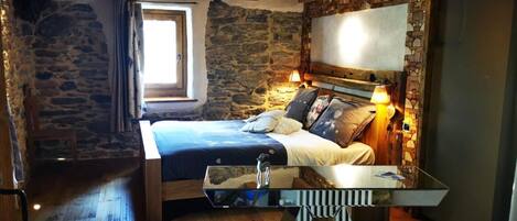 A bedroom with a stone wall, wooden ceiling beams, a bed with multiple pillows, bedside lamps, and a large mirror.