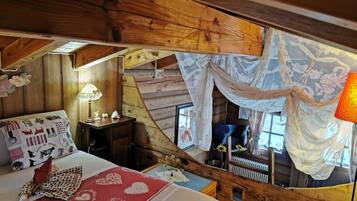 Ein gemütliches Blockhauszimmer mit Bett, Nachttisch, Lampe und einem Fenster mit transparenten Vorhängen.
