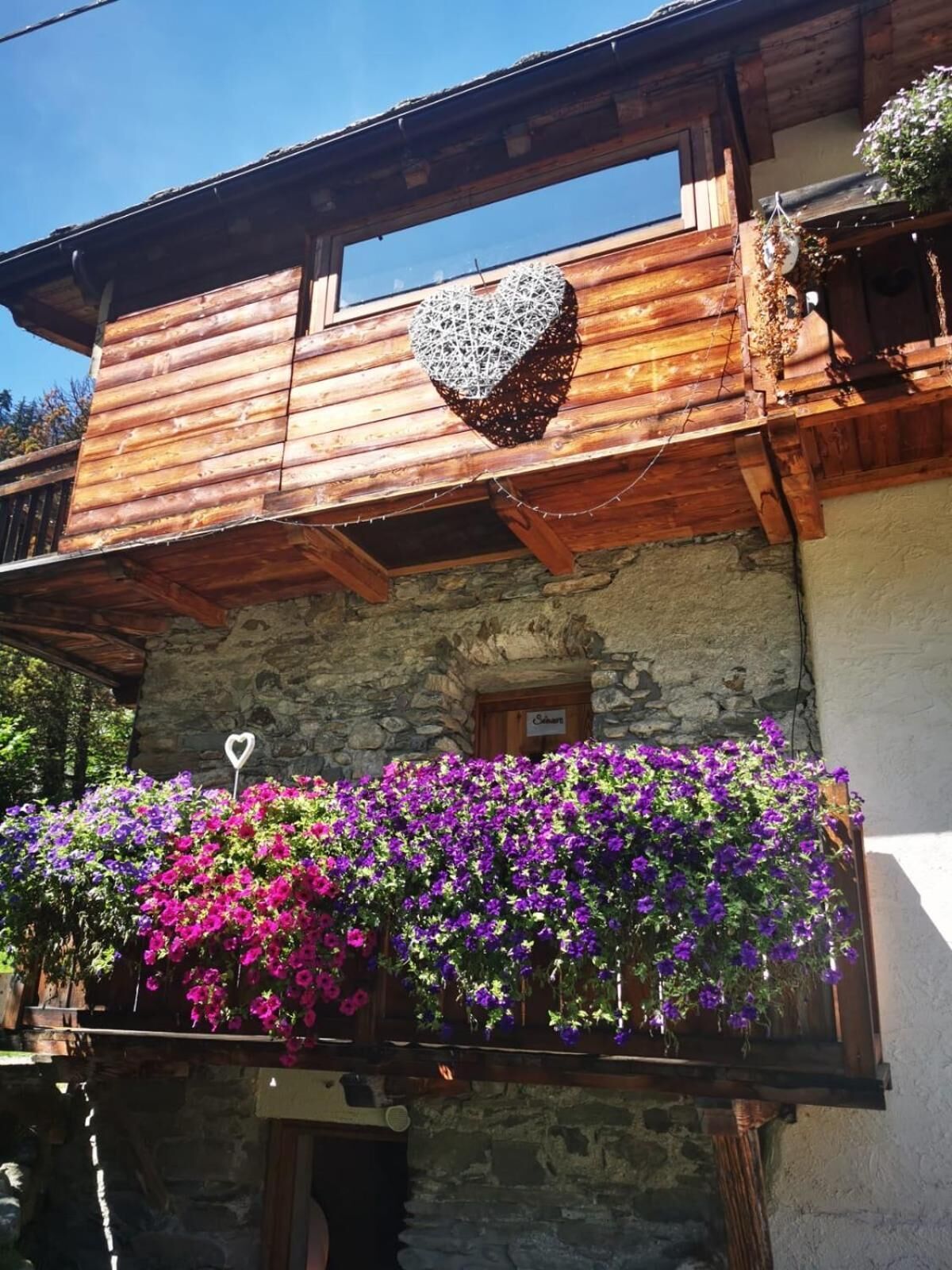 Un edificio de madera con fachada de piedra, un balcón con flores moradas y una decoración con forma de corazón.