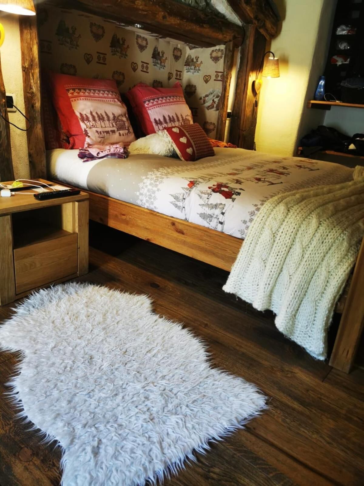 Un dormitorio acogedor con una cama de madera, ropa de cama estampada, una alfombra blanca y mullida, y una estantería con objetos personales.
