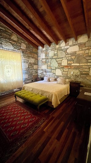 Un dormitorio con pared de piedra, vigas de madera, una cama con almohadas, un banco verde y un tapete rojo con estampado.