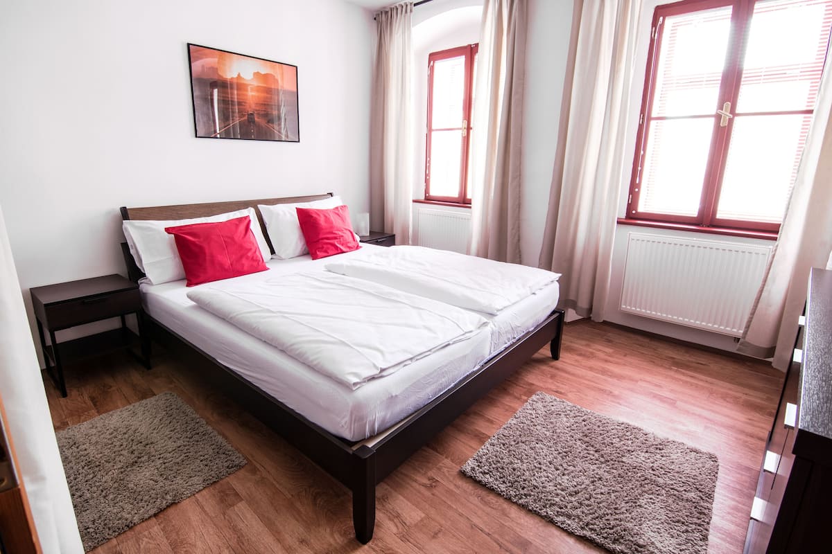A bedroom with a wooden bed, two bedside tables, a nightstand, a window with curtains, and a radiator.