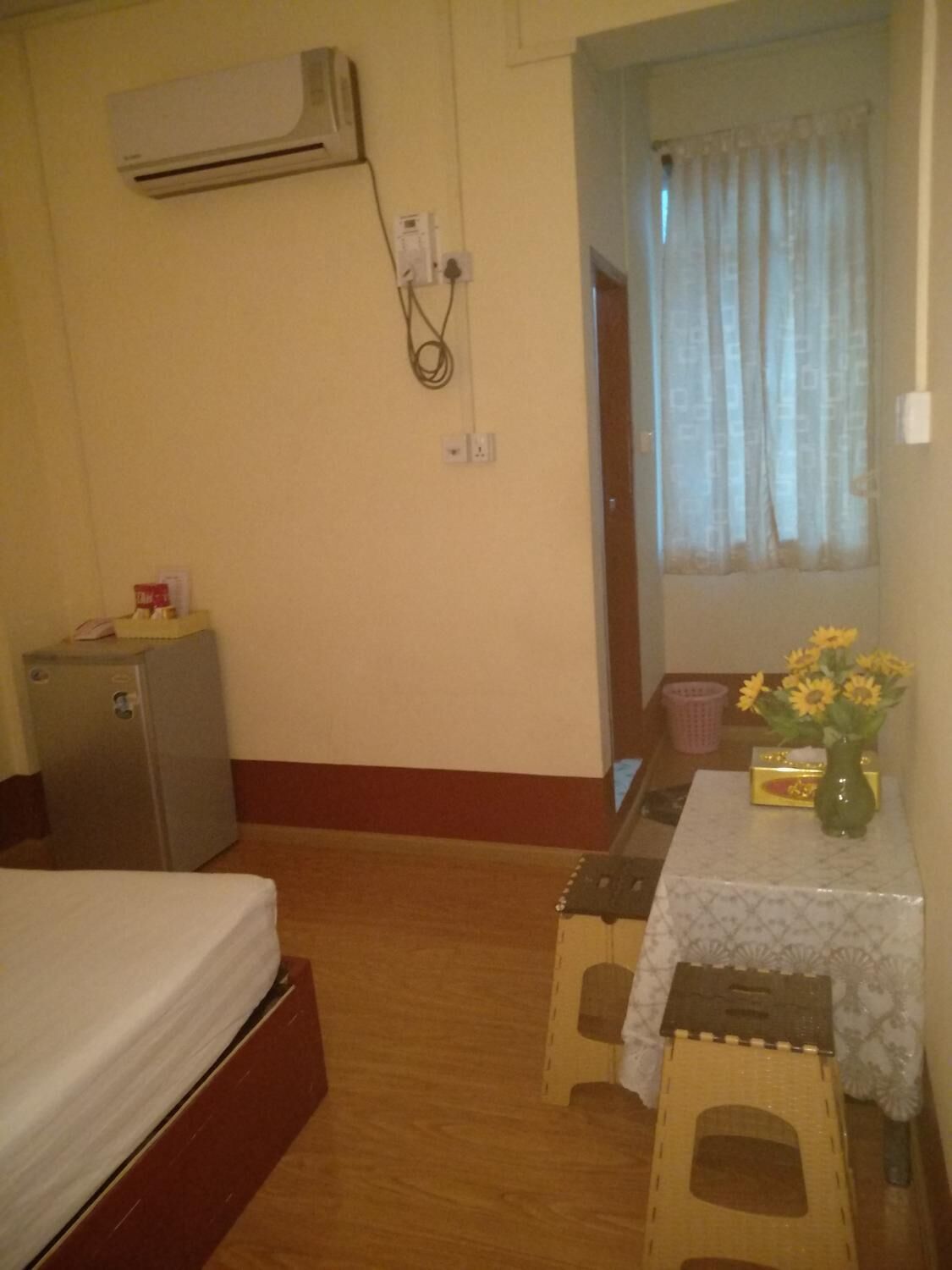 A small room with a bed, a desk with a vase of flowers, and an air conditioning unit on the wall.