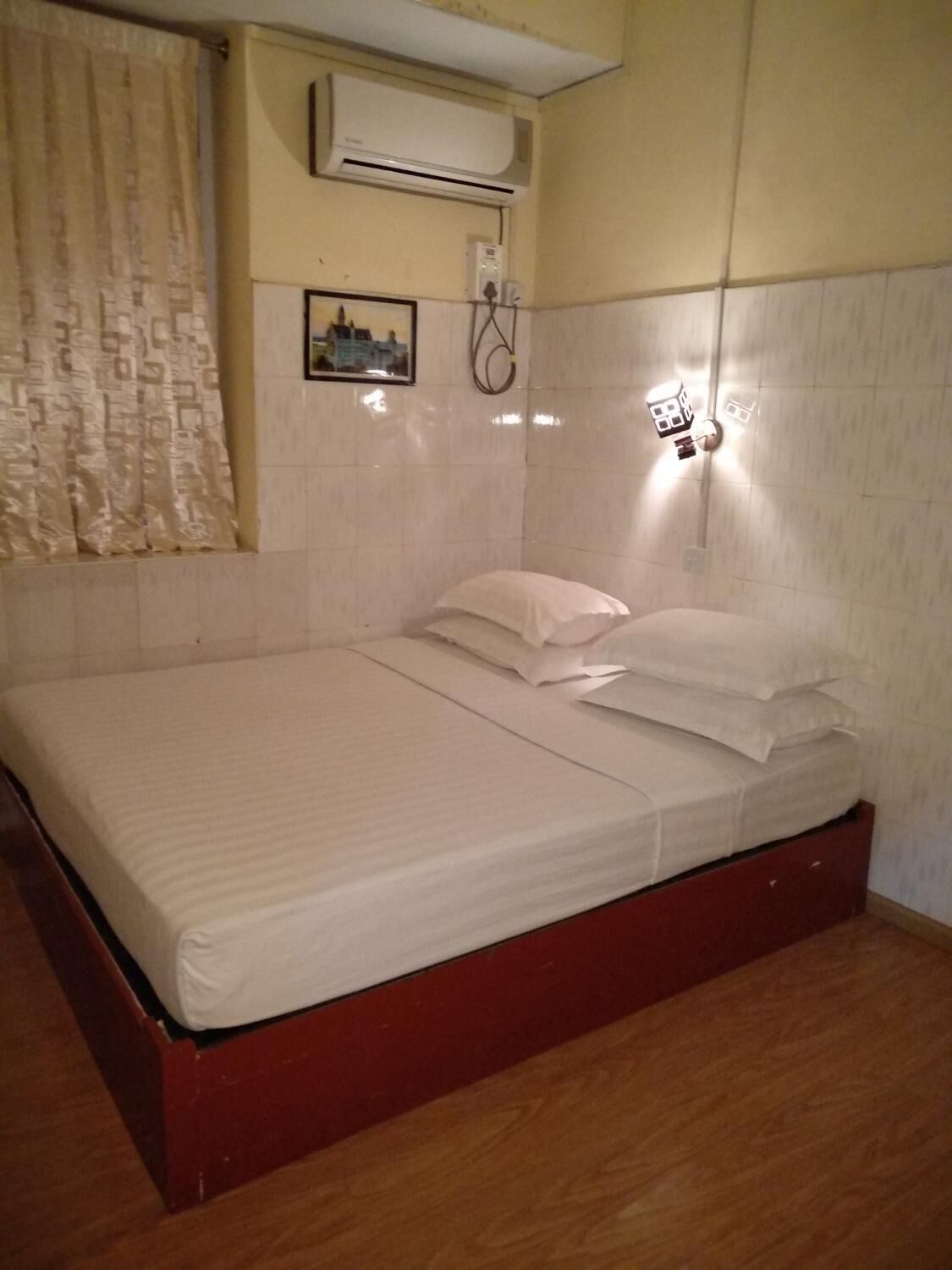 A single-bed room with a wooden headboard, a white mattress, and a red bed frame. There is a wall-mounted air conditioning unit and a small picture frame on the wall.