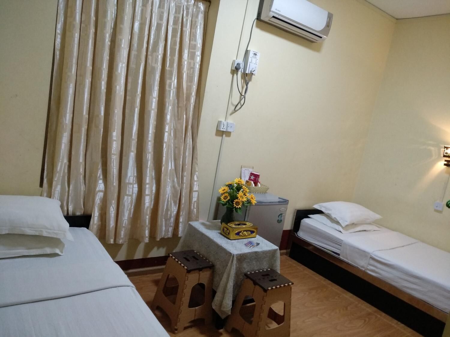 A hotel room with two beds, a small table with a vase of sunflowers, a desk with a computer, and an air conditioning unit on the wall.