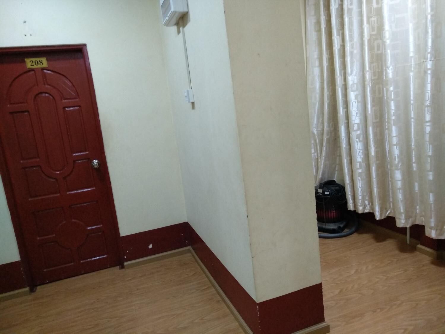 A room with a red door marked 208, a white wall, a wooden floor, and a white curtain.