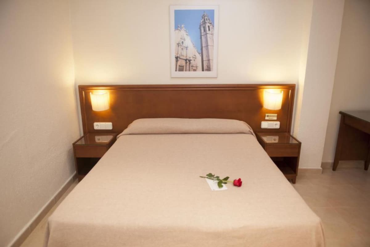 Una cama bien hecha con una sola rosa sobre el edredón, dos lámparas de noche y un cuadro en la pared.