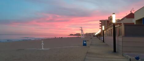 A beach with a wooden boardwalk, a lifeguard tower, and a row of buildings with lights on.
