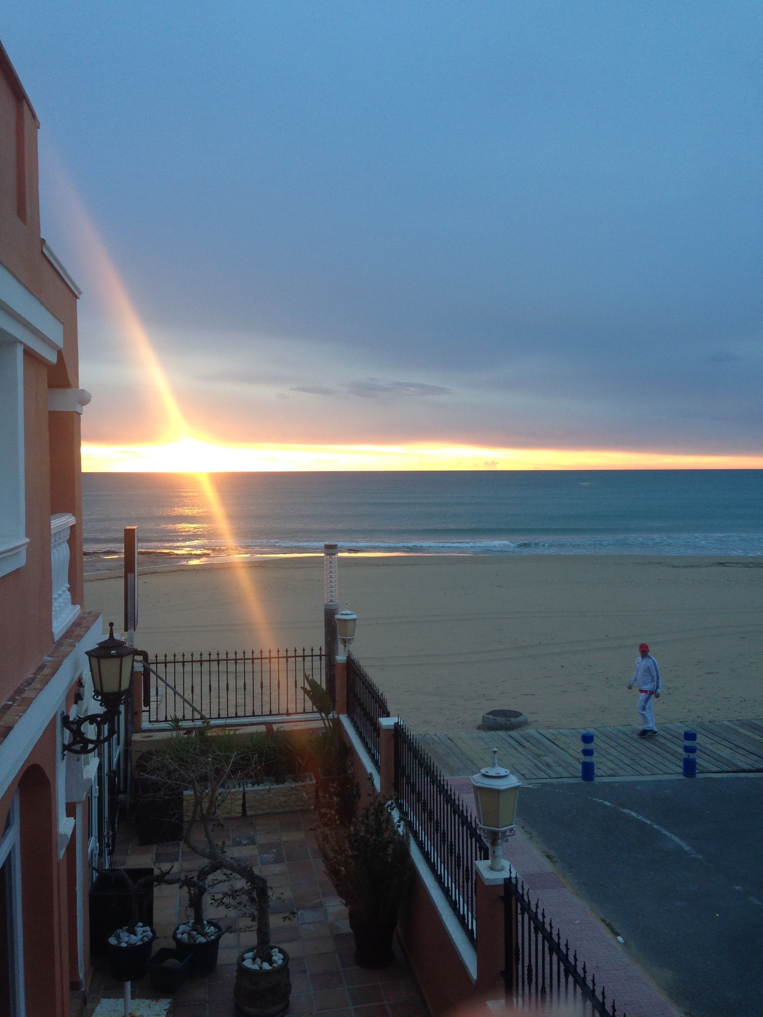 Une vue sur la côte, avec un coucher de soleil sur la plage, une personne qui se promène et un bâtiment doté d’un balcon.