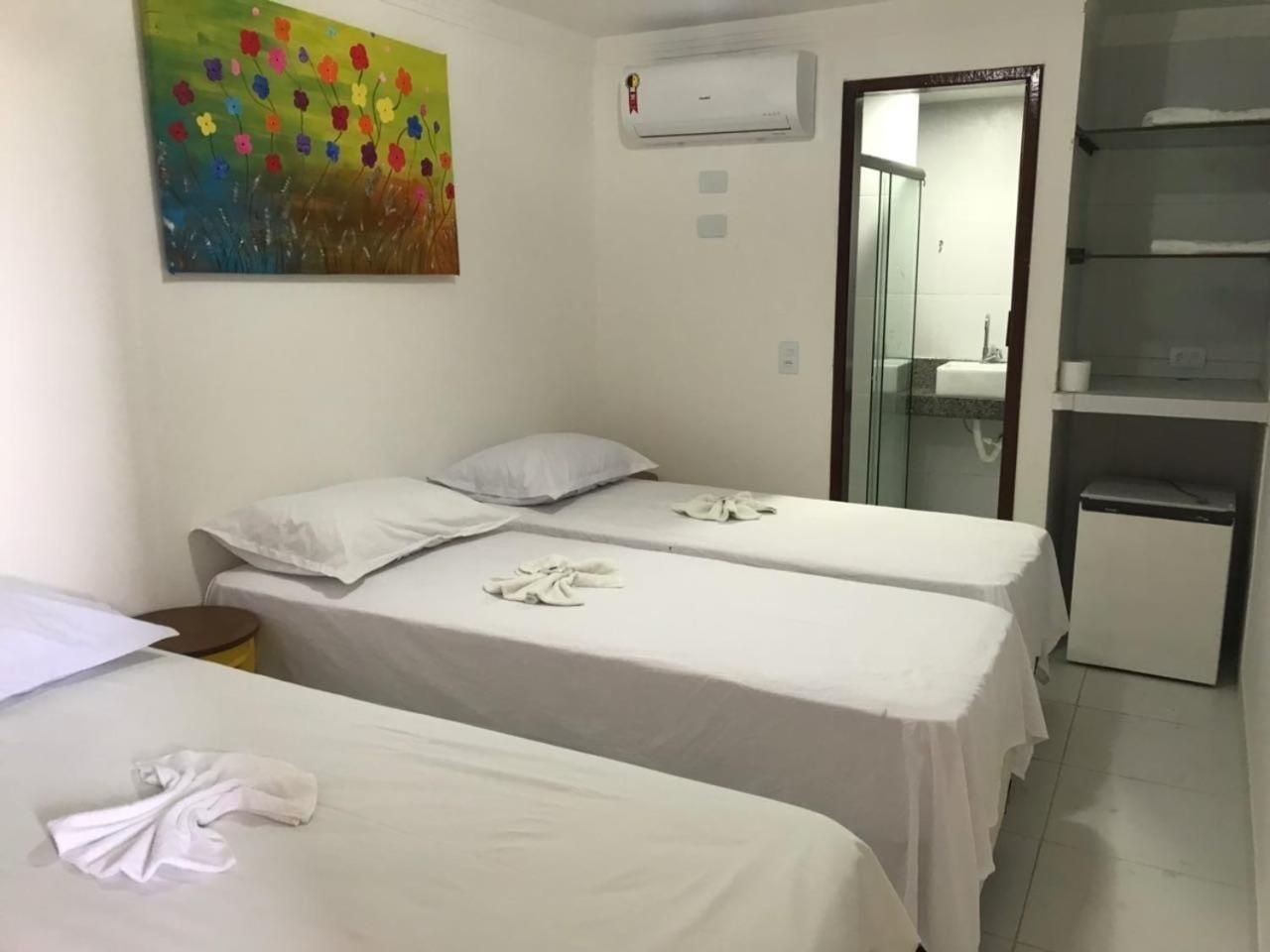 Quarto de hotel com duas camas de solteiro, uma mesinha, geladeira, ar-condicionado e um quadro colorido na parede.
