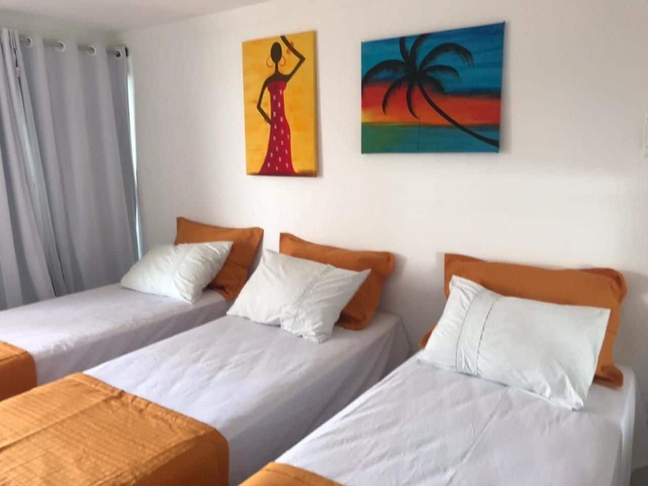 A hotel room with three single beds, each with white bedding and orange headboards. There are two paintings on the wall, one depicting a woman in a red dress and the other a tropical scene.