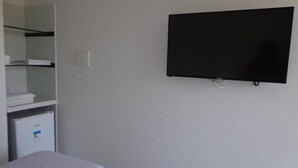 A hotel room with a bed, a TV mounted on the wall, a small fridge, and a shelf with folded towels. - Porto Palace Pousada (Ipojuca)