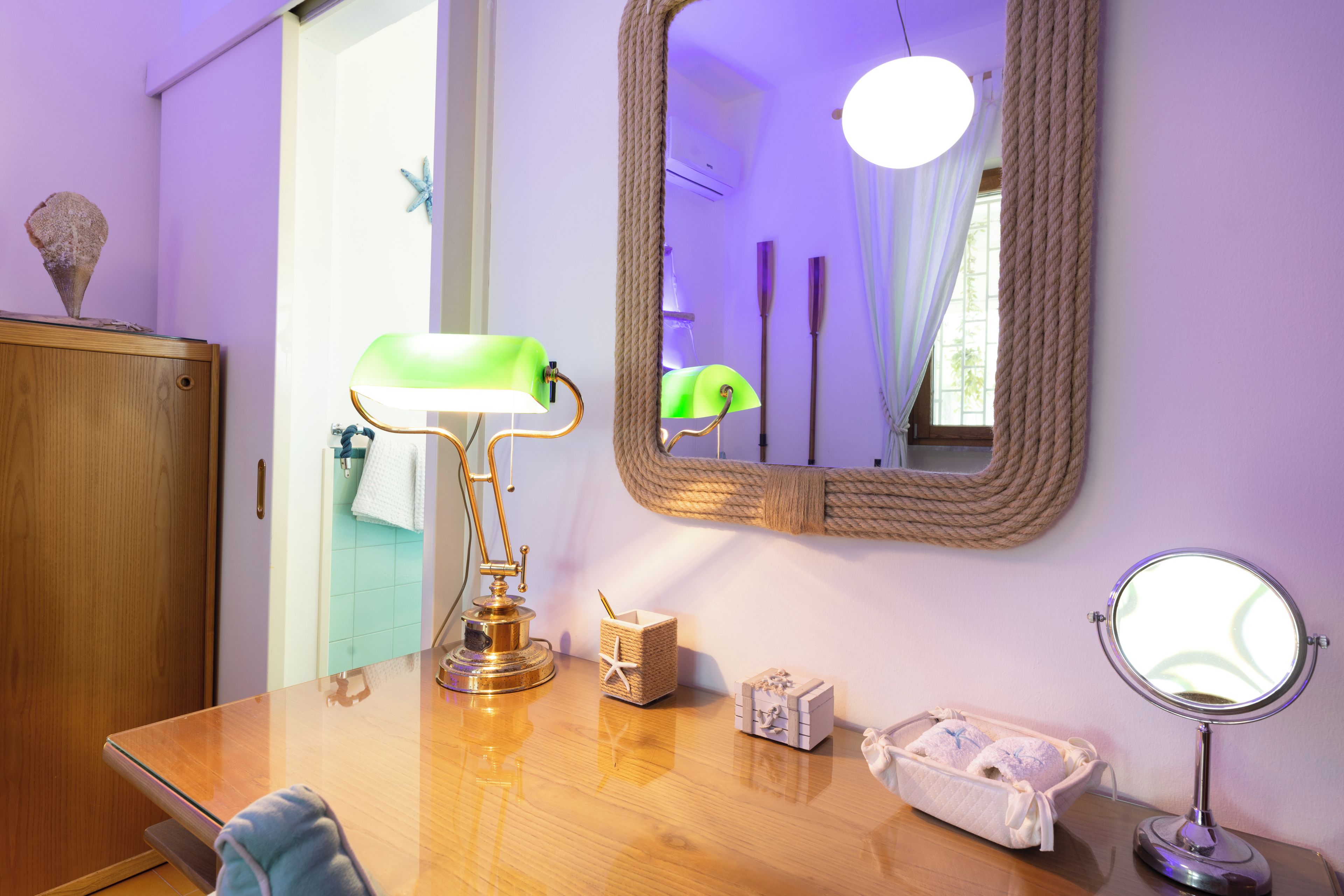 A room with a wooden desk, a green desk lamp, a mirror, and a small wooden chest.