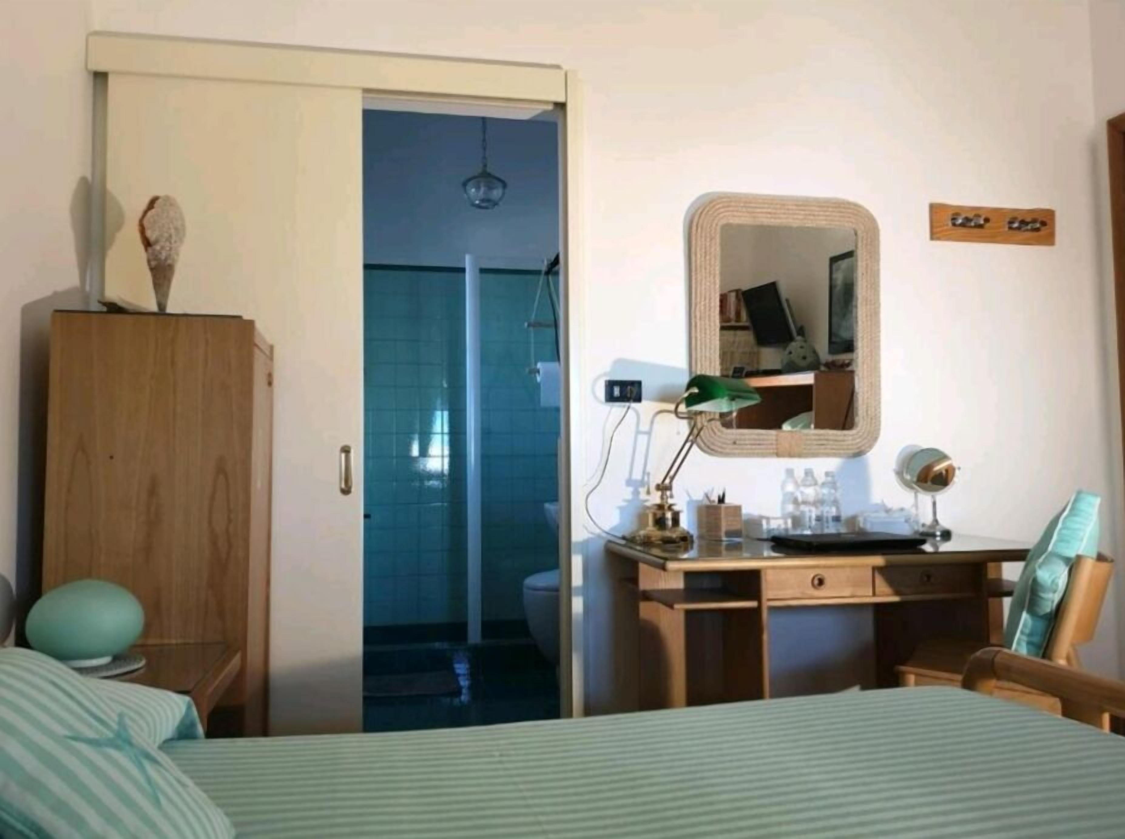 Un dormitorio con cama, armario de madera, escritorio con espejo y baño a la vista a través de una puerta abierta.
