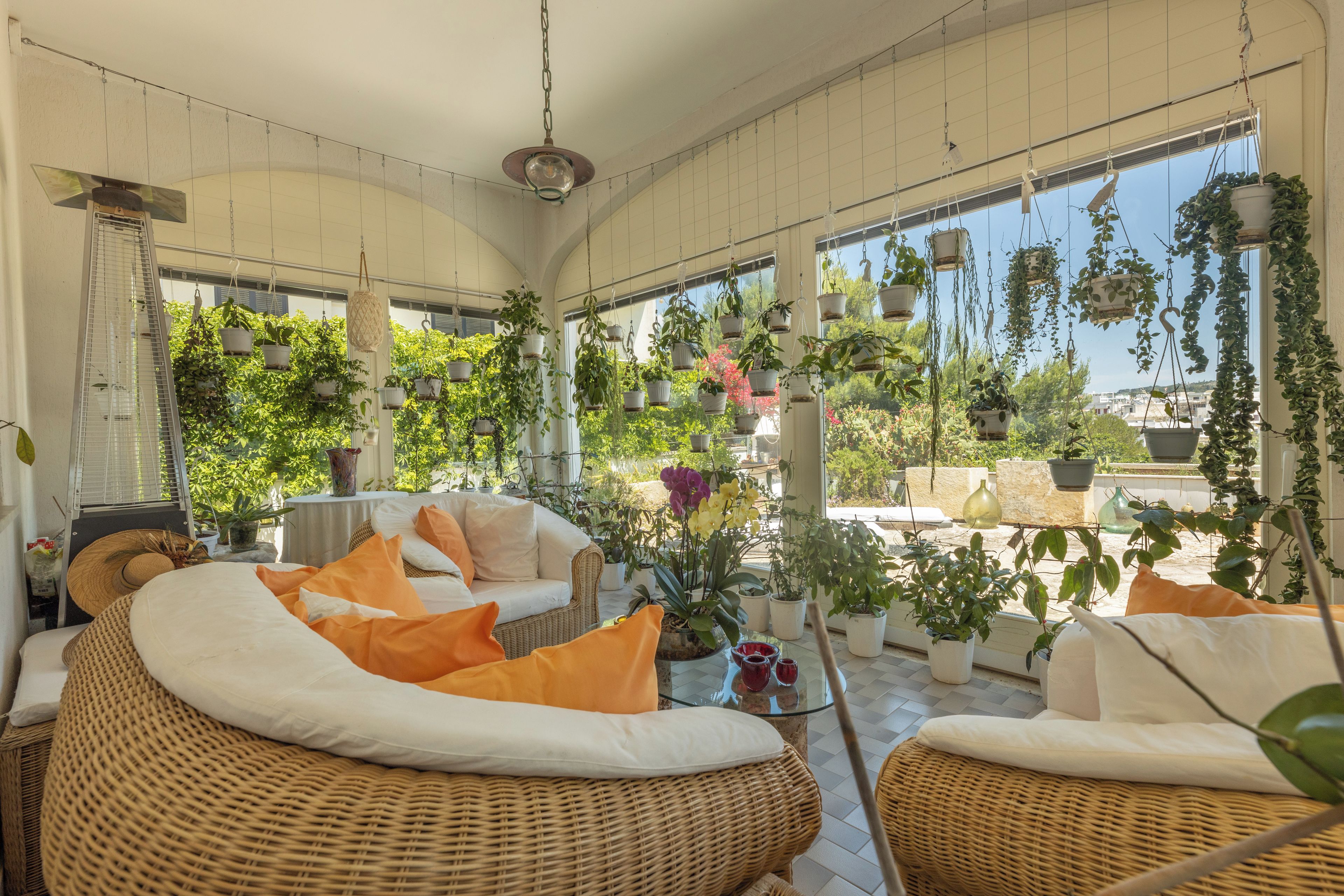 Una habitación iluminada por el sol con muebles de mimbre, una mesa de cristal y abundantes plantas colgantes.