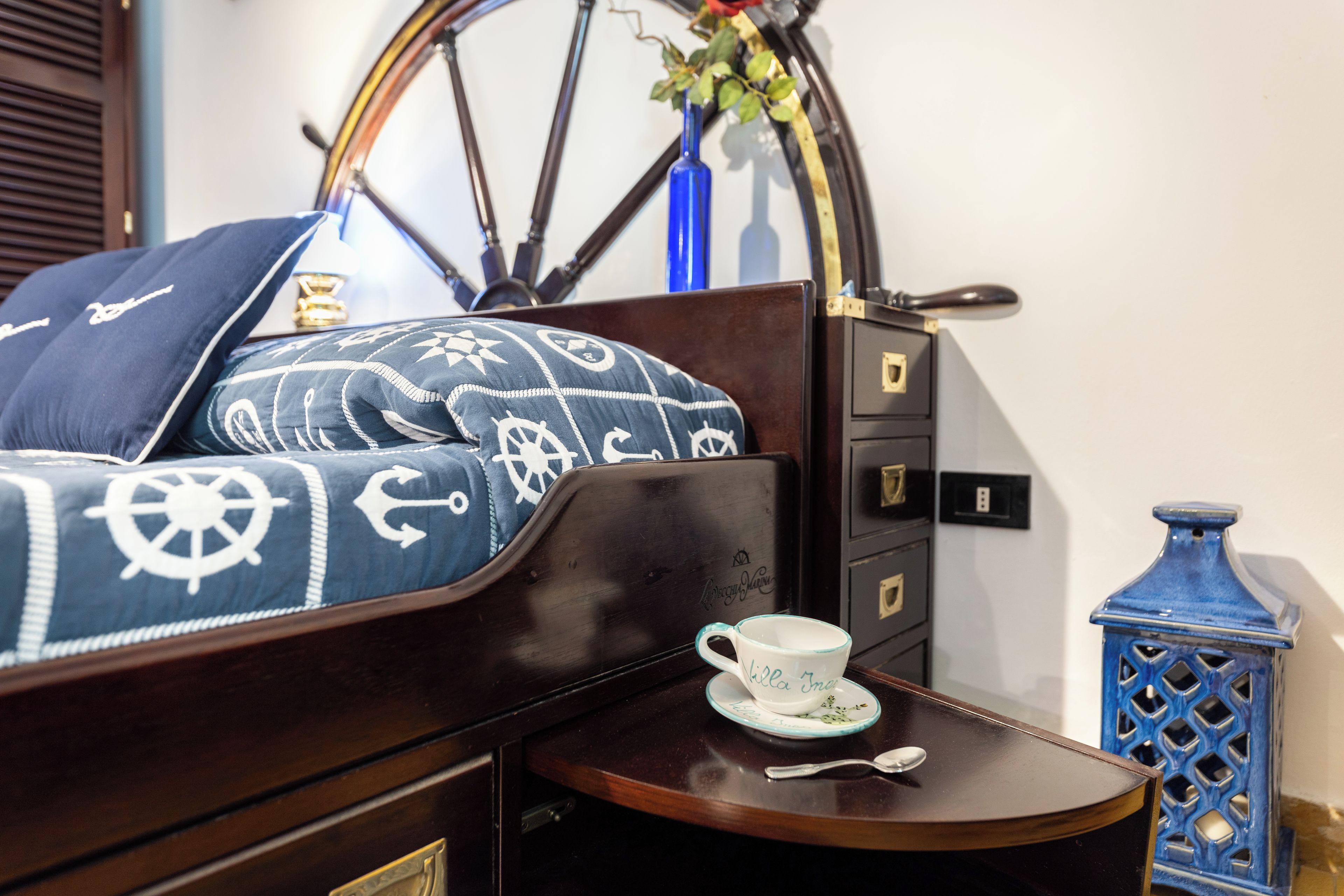 Una cama bien hecha con ropa de cama estampada, una mesita de noche con una taza y una cuchara, un jarrón azul con flores y un aparador de madera con cajones.