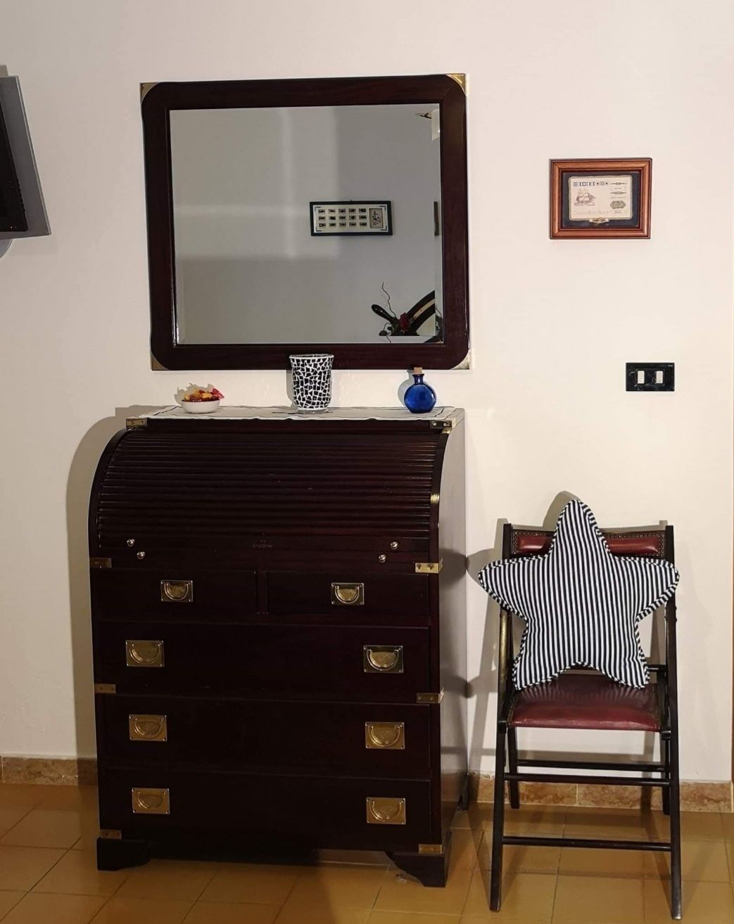 Una habitación con un aparador de madera oscura, una mesita con un jarrón azul, una silla con un cojín a rayas, un espejo grande y un diploma enmarcado en la pared.