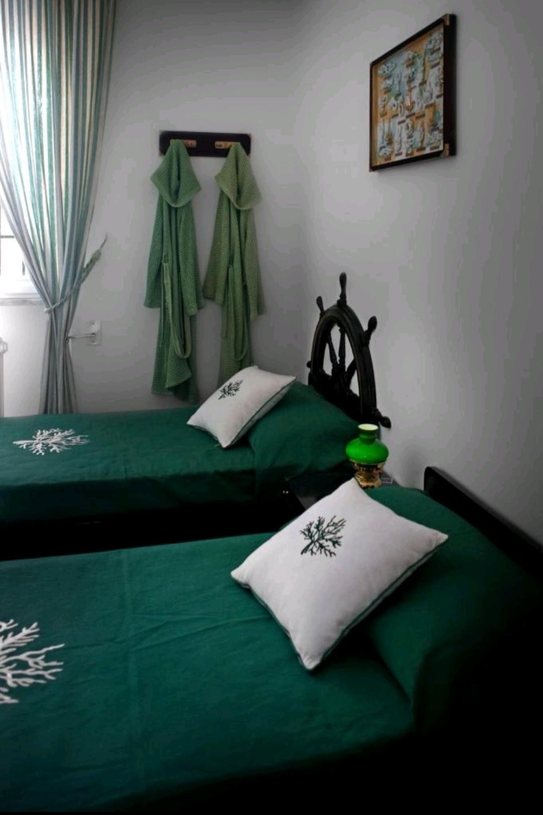 A room with two beds, each with a green bedspread and a white pillow with a tree design. There are green towels hanging on a rack, a ship's wheel decoration on the wall, and a framed picture above the beds.