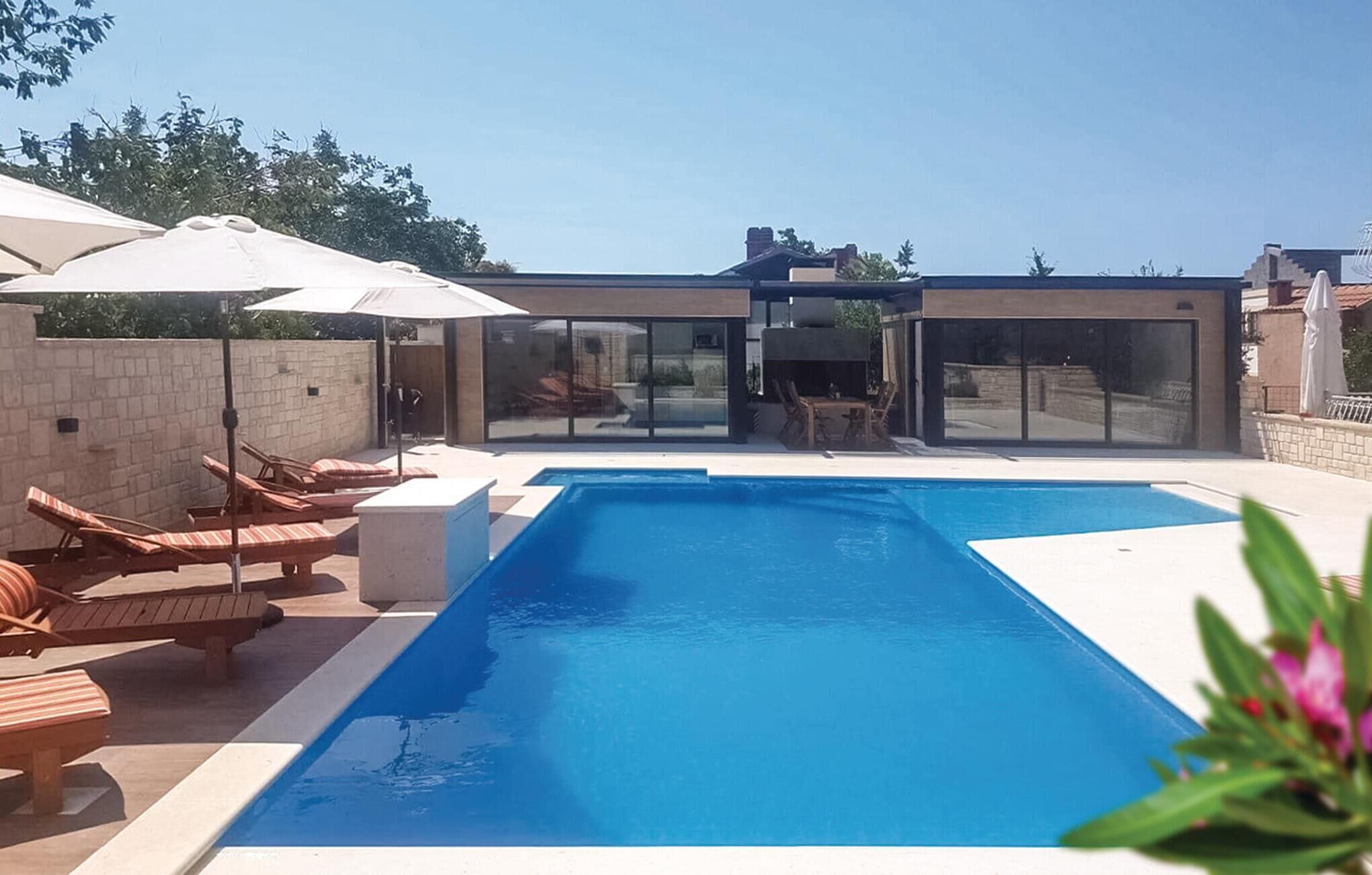 Ein moderner Außenpool mit Liegestühlen und Sonnenschirmen, umgeben von einer Steinmauer und Glastüren, die zu einer überdachten Terrasse führen.