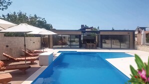 Una piscina moderna al aire libre con sillas de descanso y sombrillas, rodeada por un muro de piedra y puertas de cristal que dan a un porche cubierto.