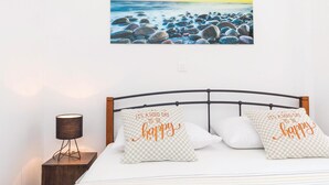 Un dormitorio con cama, dos almohadas con el texto "It's a good day to be happy", una mesita de noche con lámpara y un cuadro de una playa rocosa.