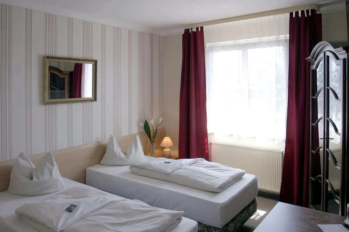 A hotel room with two beds, a mirror, a window with red curtains, and a radiator.