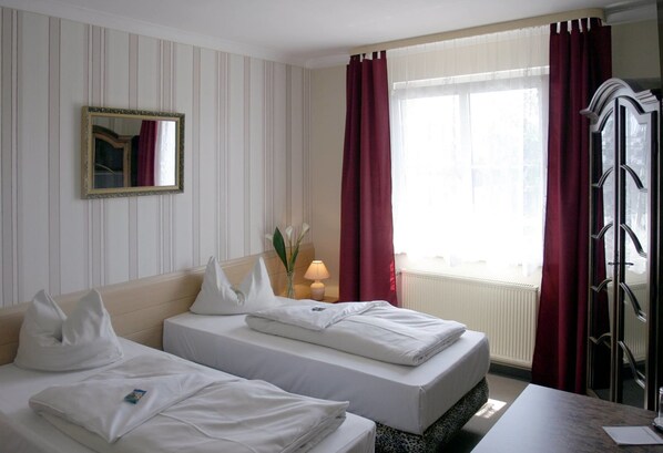 A hotel room with two beds, a mirror, a window with red curtains, and a radiator.