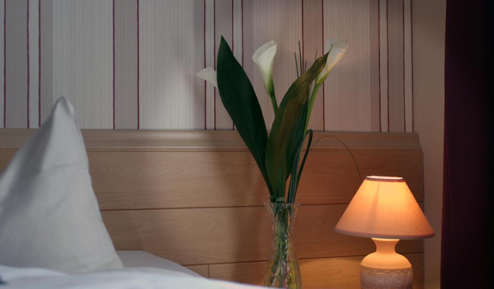 A hotel room with a wooden headboard, a bedside table with a lamp, and a vase with flowers.