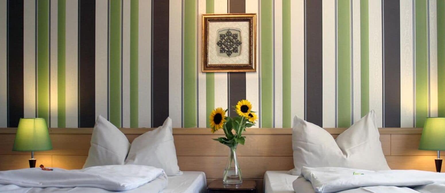 Ein Hotelzimmer mit gestreifter Tapete, zwei Betten mit weißen Leinentüchern, einer Vase mit Sonnenblumen und einem gerahmten Bild an der Wand.