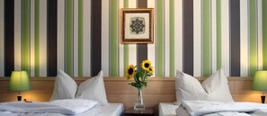 A hotel room with striped wallpaper, two beds with white linens, a vase with sunflowers, and a framed picture on the wall.