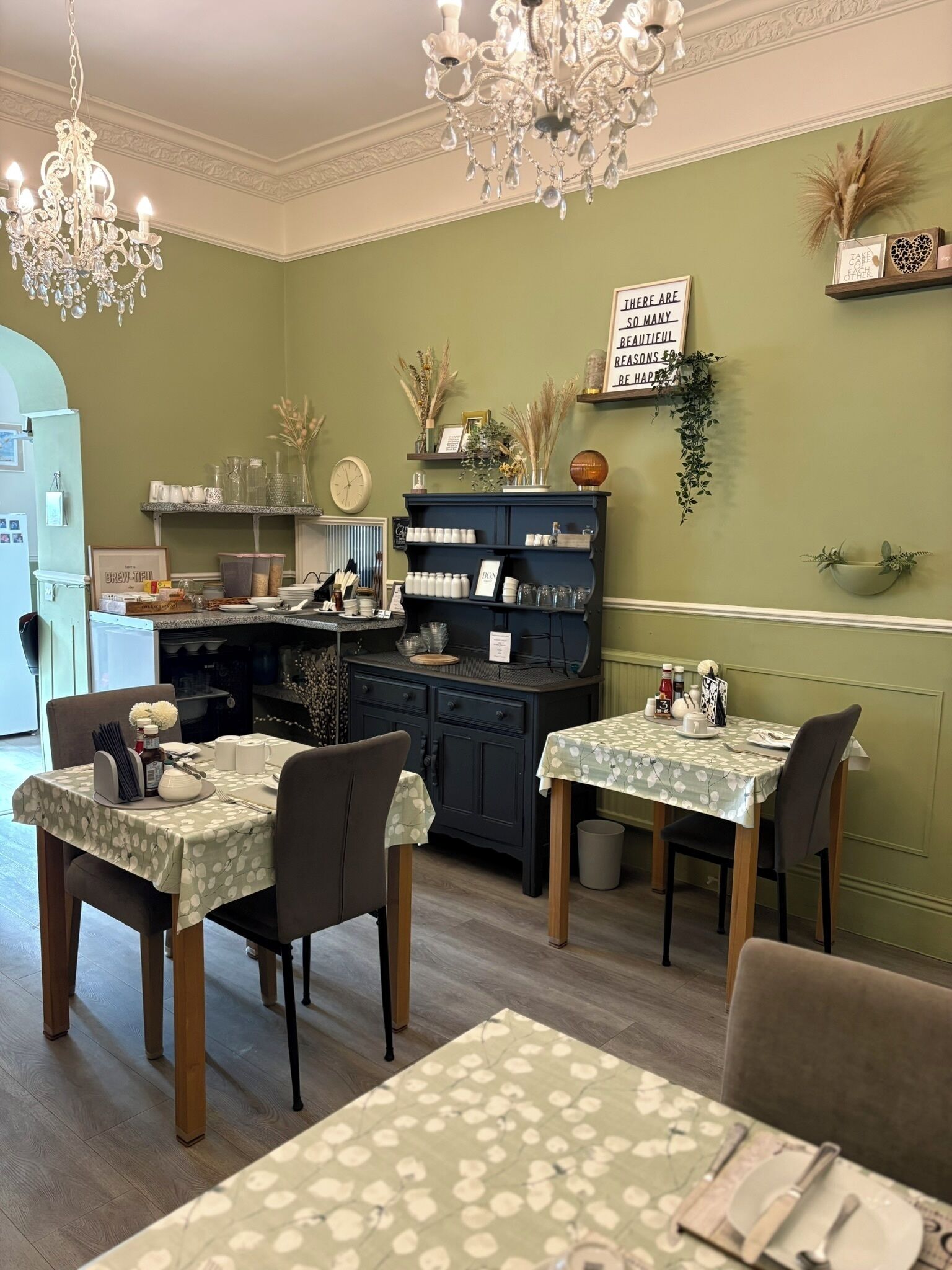 A dining area with two tables, chairs, a chandelier, and a wall-mounted shelf with decorative items.