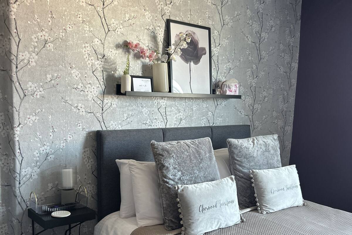Une chambre à coucher comprenant un lit, des oreillers, une table de chevet, une étagère avec des objets décoratifs et un mur orné d’un motif floral.