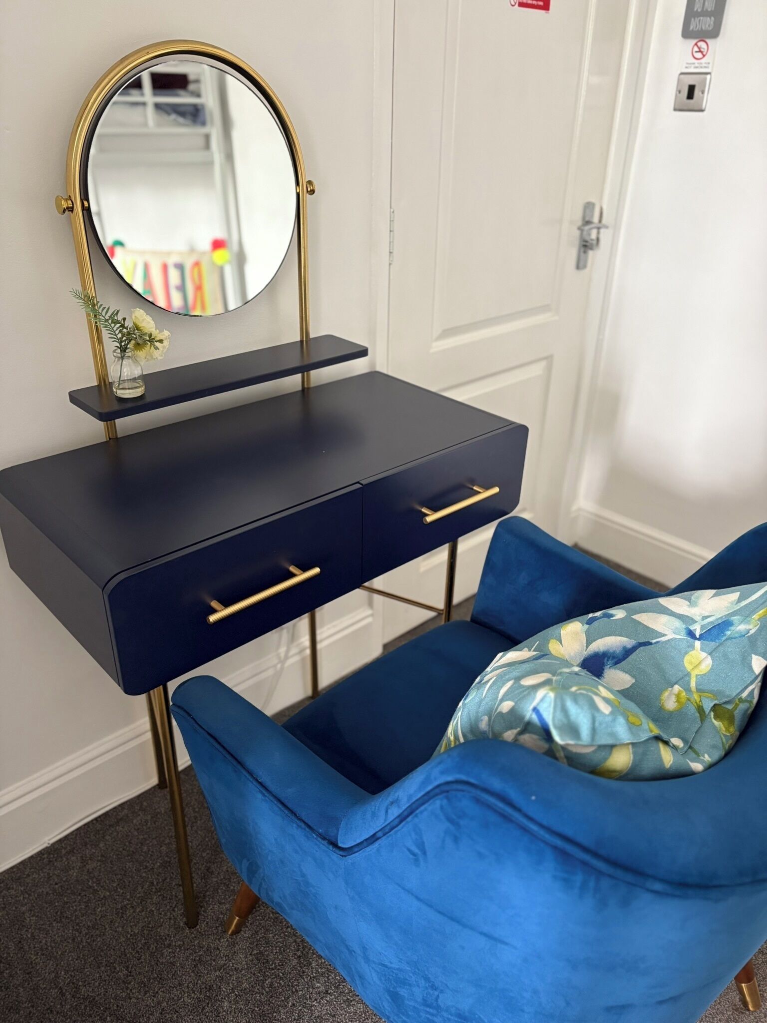 A room with a dark blue vanity with gold handles, a round mirror, and a blue chair with a floral pillow.
